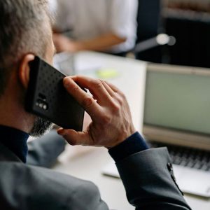 A man in a suit sits at a desk, holding a smartphone to his ear and looking at a laptop that reflects TECAR about us and international automotive expertise. The photo is taken from behind and focuses on his hand and the phone.